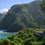 A photo of the road to Hana showing a car hugging a turn, the jungle, and the ocean below.