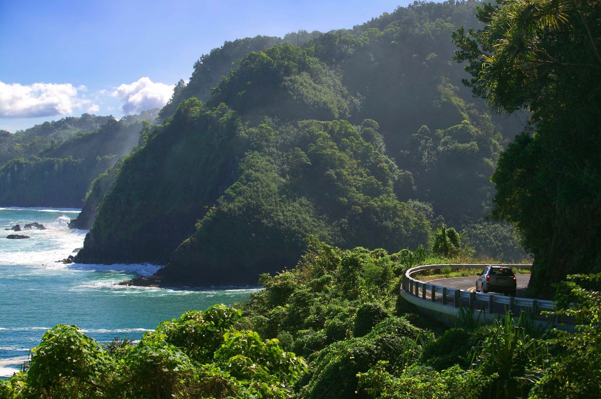 A photo of the road to Hana showing a car hugging a turn, the jungle, and the ocean below.
